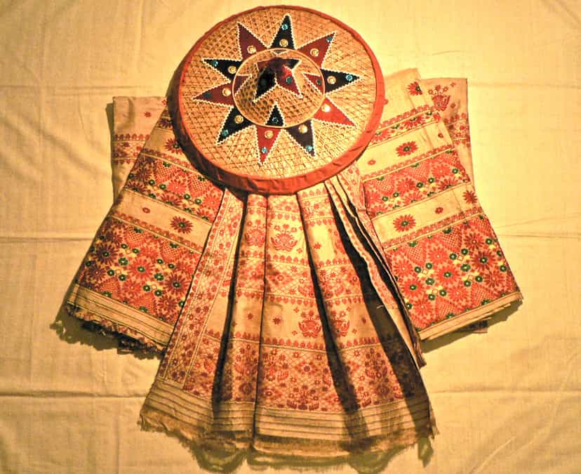 A length of golden muga silk mekhela-chador woven with red and gold floral and pictorial borders, laid out with a decorated japi