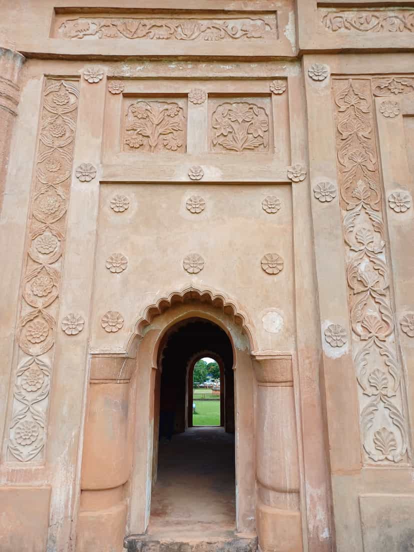 Close view of the Rang Ghar's brick wall at Sivasagar, covered in moulded terracotta ornament: bands of rosettes, framed floral relief panels, and a scalloped arched niche opening to greenery beyond