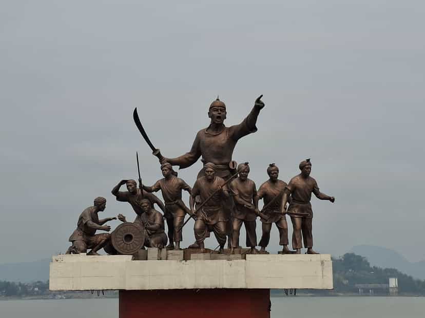 Bronze statue group: Lachit Borphukan standing tall with a raised sword and an outflung pointing arm, a band of Ahom warriors and a seated gunner with a cannon arrayed below on a white plinth, the Brahmaputra and far hills behind