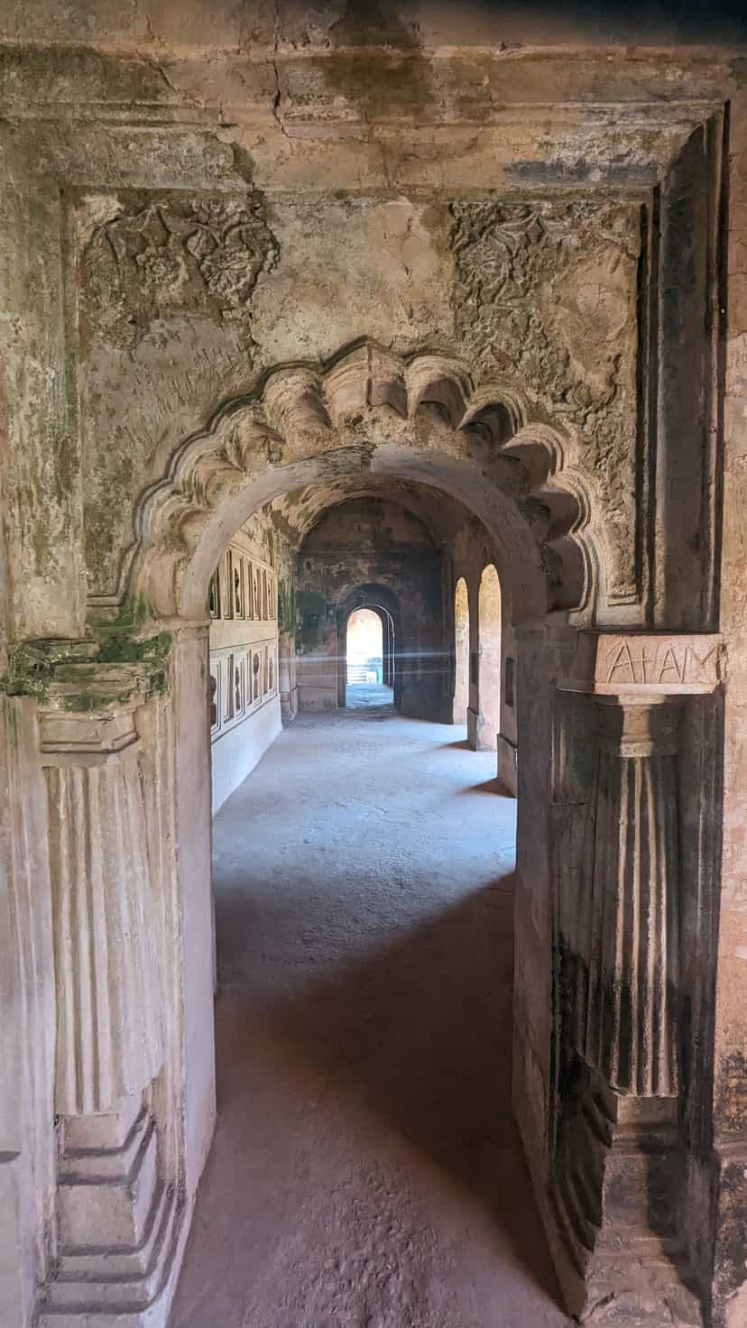 A scalloped multifoil brick arch inside the Talatal Ghar, carved plaster floral spandrels above it and a second arched hall with niched walls visible beyond, leading to a small lit opening at the far end