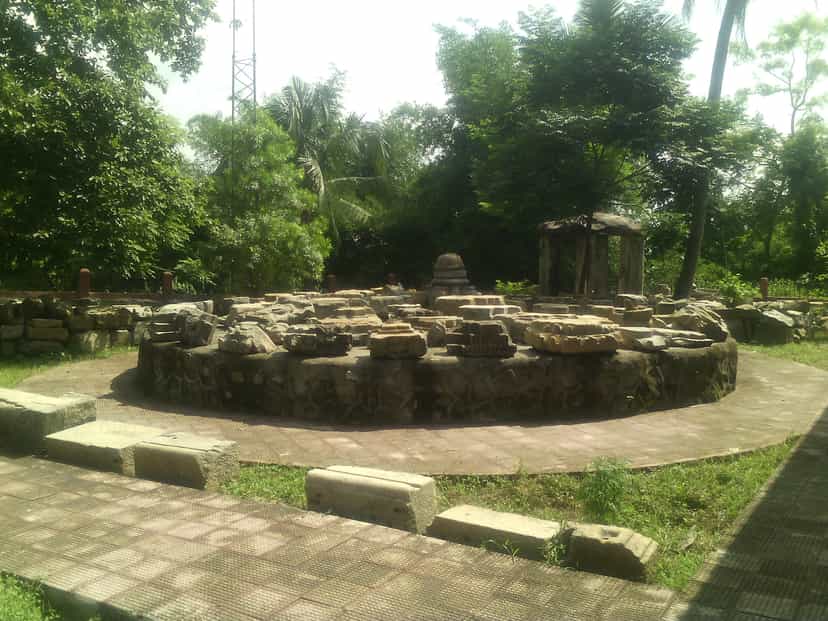 Circular stone plinth and scattered carved blocks of the ruined Madan Kamdev temple complex in a landscaped clearing