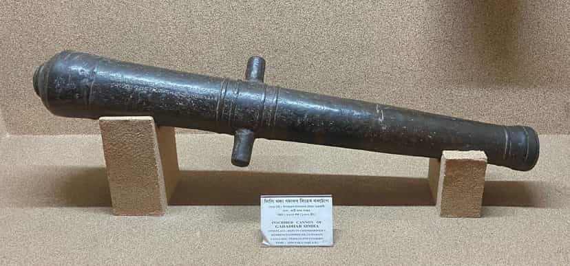 An old iron cannon resting on two brick supports in a museum display, its body bearing an Assamese inscription, with a label reading inscribed cannon of Gadadhar Simha