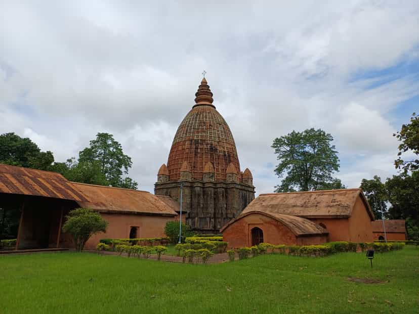A tall brick temple with a ribbed, curvilinear spire rising over lower red-brick outbuildings with curved roofs, set behind a mown lawn and hedge under a cloudy sky