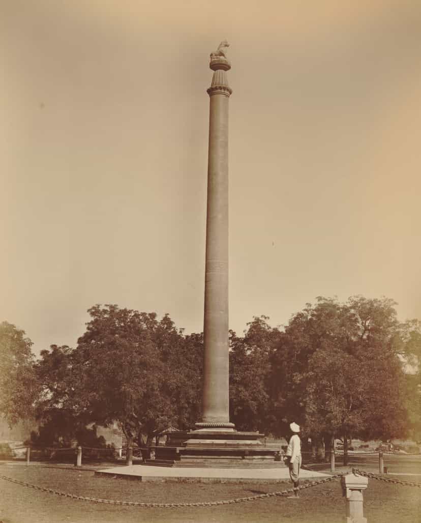 A tall polished sandstone column standing on a stepped plinth among trees, a small figure at its base for scale