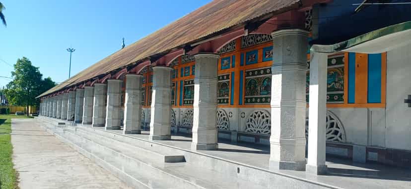 The long pillared veranda of the Barpeta Sattra kirtan-ghar in bright sunlight, a row of stout whitewashed columns on a raised plinth receding in perspective beneath a low pitched roof, the wall behind painted in orange with figural panels and pierced white lattice screens, under a clear blue sky