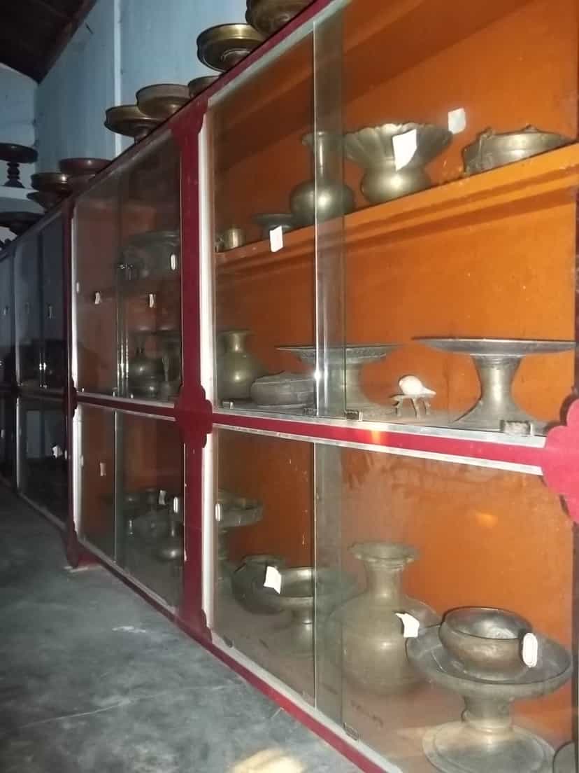 Glass display cases of brass and metal ritual vessels — bowls, footed trays, water pots — at Bengenaati Sattra