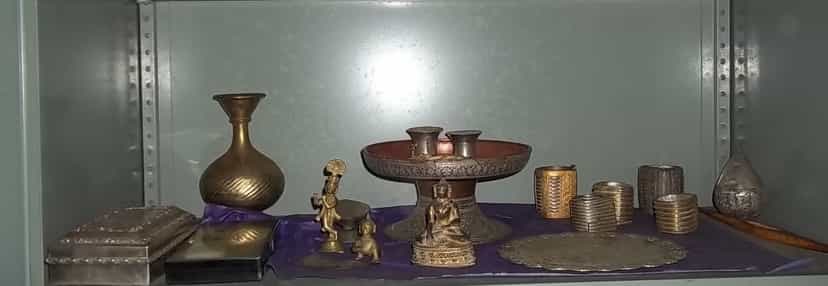 A shelf of gold and silver objects — a gilt Krishna figure, an ornate offering stand, silver caskets and bangles — at Bengenaati Sattra