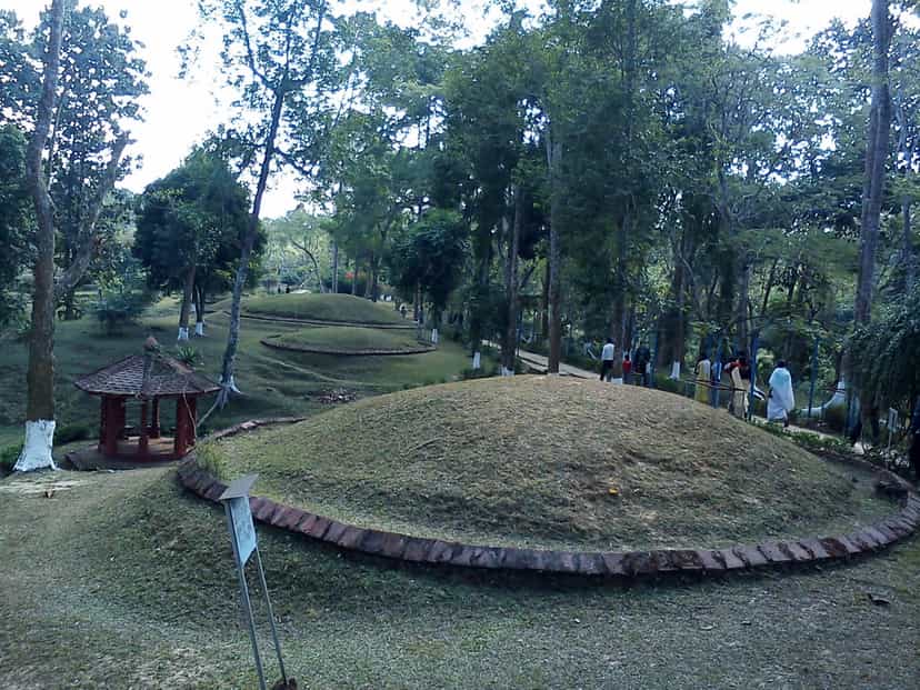A grass-covered Ahom maidam at Charaideo ringed at its base by a low octagonal wall of red brick, a small white pavilion beside it and further green mounds rising along the wooded ridge behind