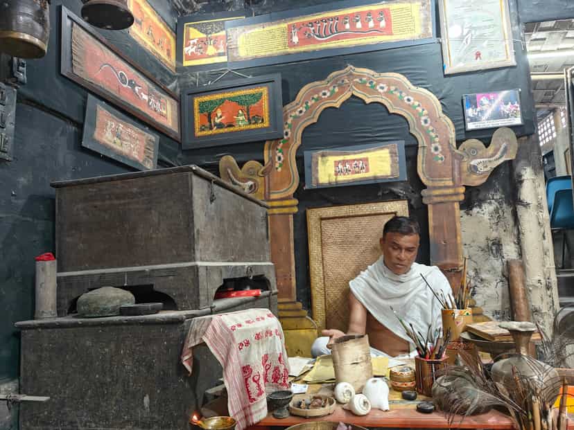 A sattra artist seated beneath a carved and gilded niche hung with painted manuscript folios, working with traditional materials at Garamur on Majuli