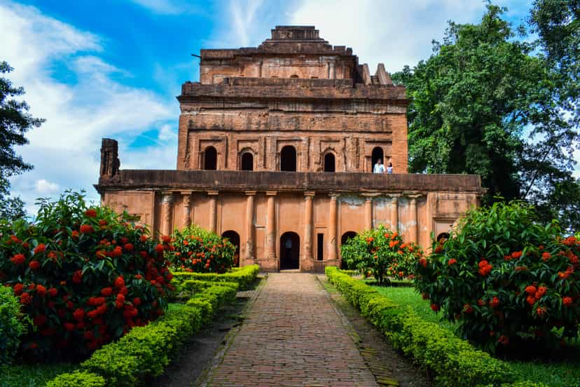 The Kareng Ghar palace at Garhgaon — a tall multi-storeyed Ahom brick palace whose upper floors step back as they rise, approached by a garden path between flowering shrubs