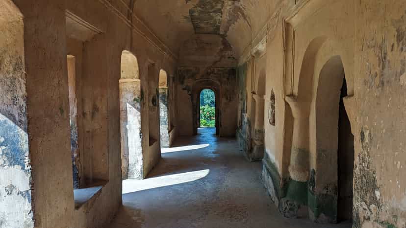 A long barrel-vaulted gallery of a ruined masonry palace, its plaster weathered and green-stained, with thick piers and recessed arched niches running down both walls, sunlight falling across the bare floor, and a small arched doorway at the far end opening onto greenery