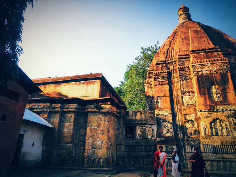 The laterite-red stone Hayagriva Madhava temple at Hajo in warm evening light, its curved carved shikhara tower beside the lower hall, framed by green trees under a deep blue sky