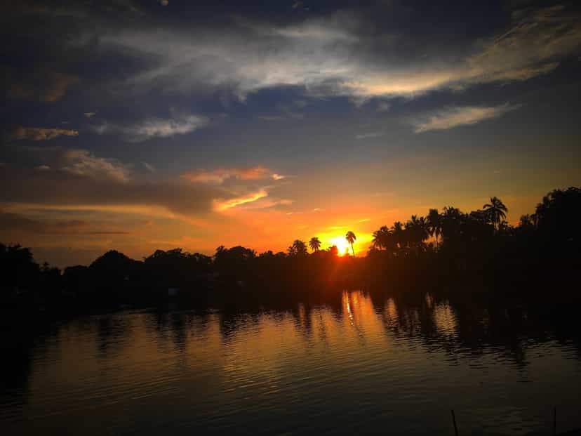 A vivid orange sunset low over a still tank at Hajo, dramatic streaked clouds overhead and palms silhouetted along the far bank reflected in the water