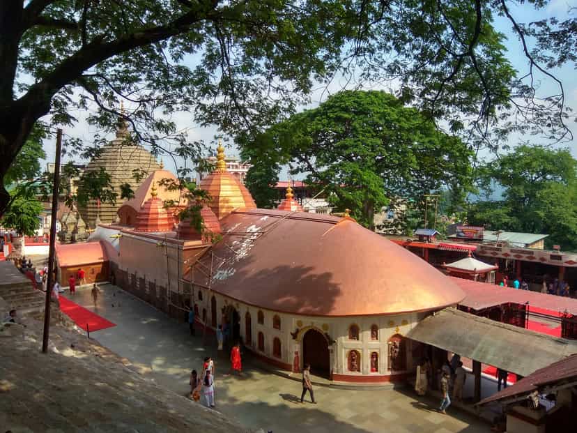Elevated daytime view of the Kamakhya temple complex: the curved copper-coloured roof of the natamandira in front, the beehive shikhara and tiered red turrets rising behind, framed by green trees