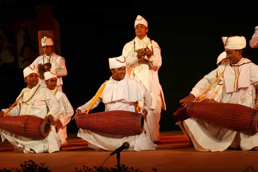 Men in white dress and white turbans kneeling and playing the khol drum, with a cymbal player standing, on a performance stage
