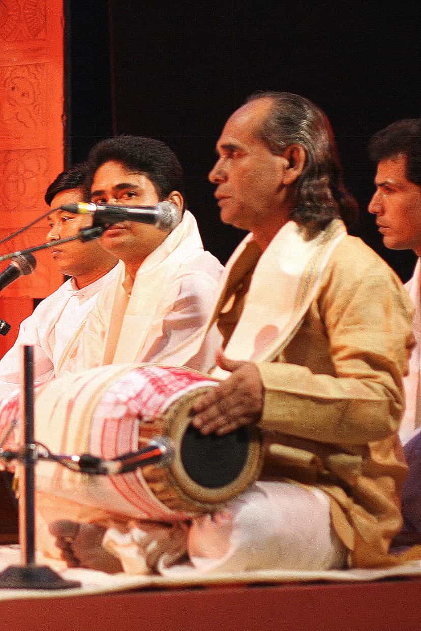 An older man in cream dress with a khol drum across his lap, singing among seated musicians at microphones on a stage