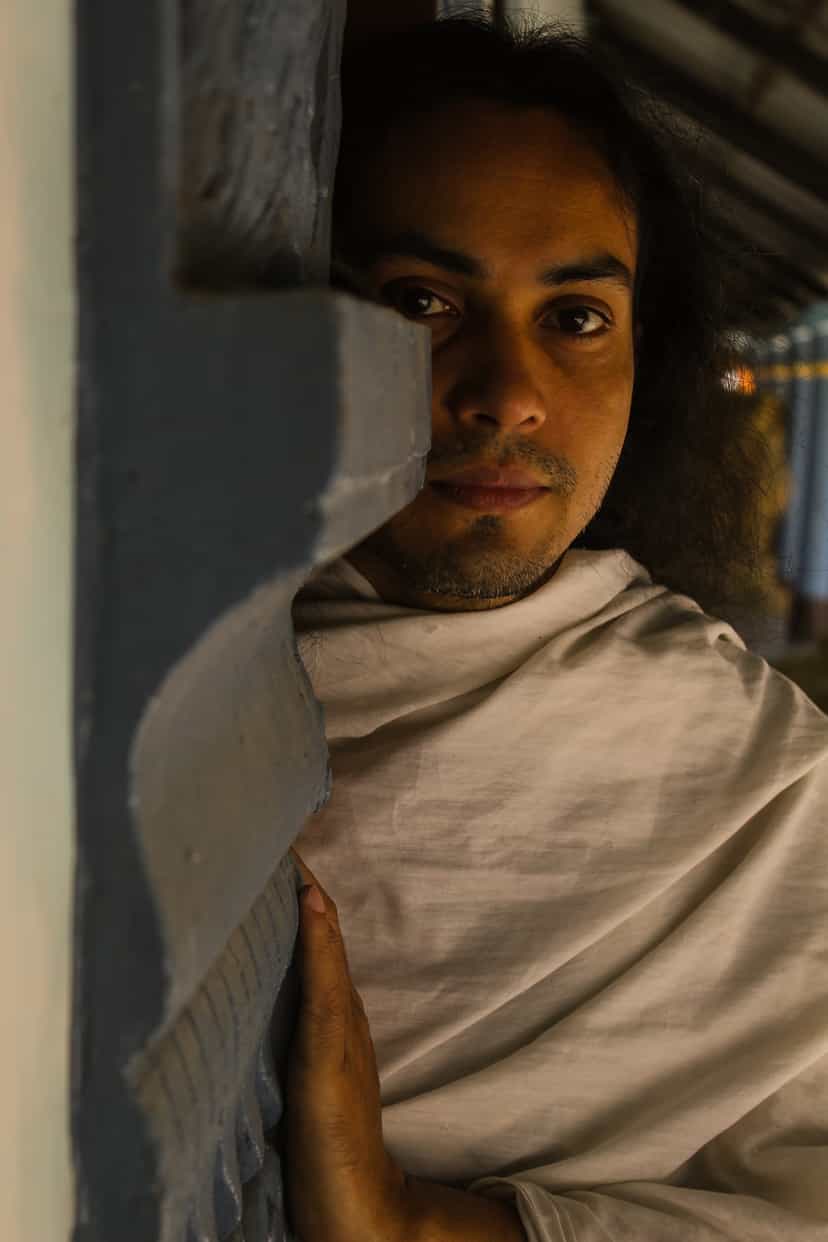 A young monk wrapped in a plain white cloth leans against a painted blue doorframe of the monastery, lit by warm low-angle light, looking calmly toward the camera