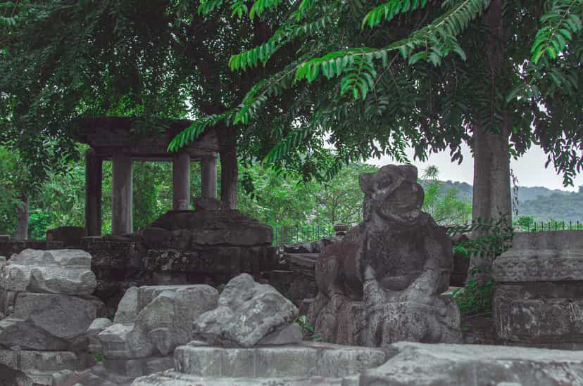 The ruins of Madan Kamdev on its wooded hill: a pillared stone shrine remnant at left, a carved guardian-lion sculpture on a plinth at right, and scattered carved stone blocks in the foreground, framed by an overhanging tree with the low hills beyond