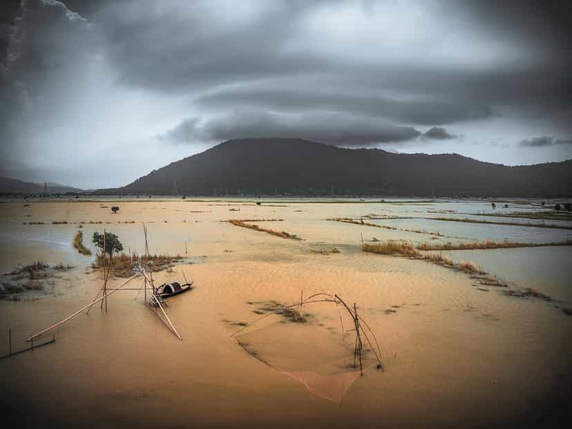 Amber floodwater spreading across Pobitora's grassland below a hill under a heavy monsoon sky, with a small boat and fishing poles