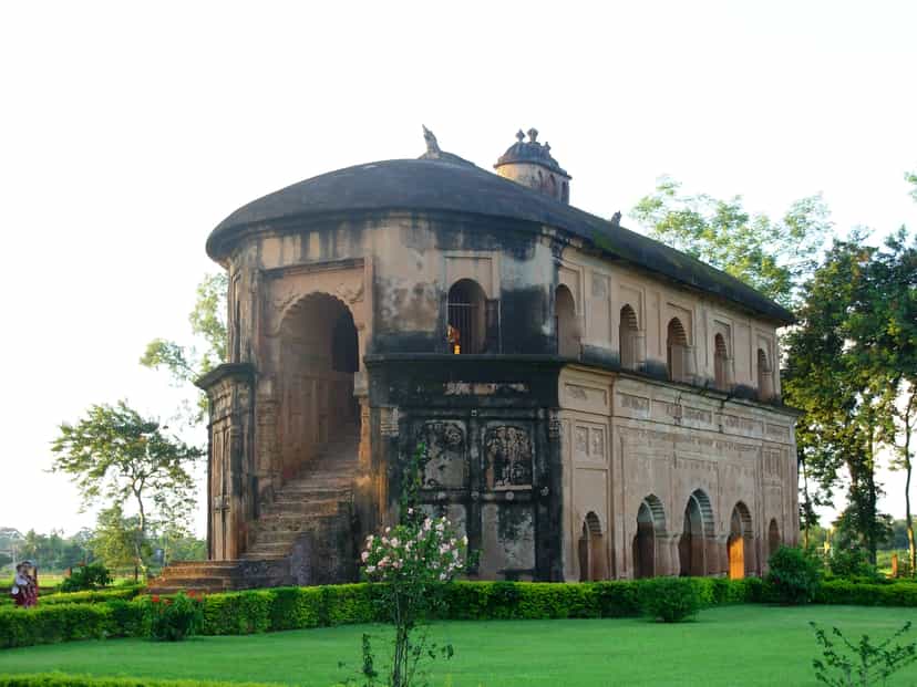 Rear three-quarter view of the Rang Ghar in warm afternoon light, showing the external masonry ramp that climbs to the upper gallery, with carved panels on the curved end and green surroundings