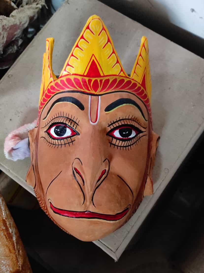A monkey-faced Hanuman mukha resting on a plain surface, its long pink-and-ochre muzzle and wide painted eyes below a yellow-and-red pointed crown, a Majuli bhaona mask
