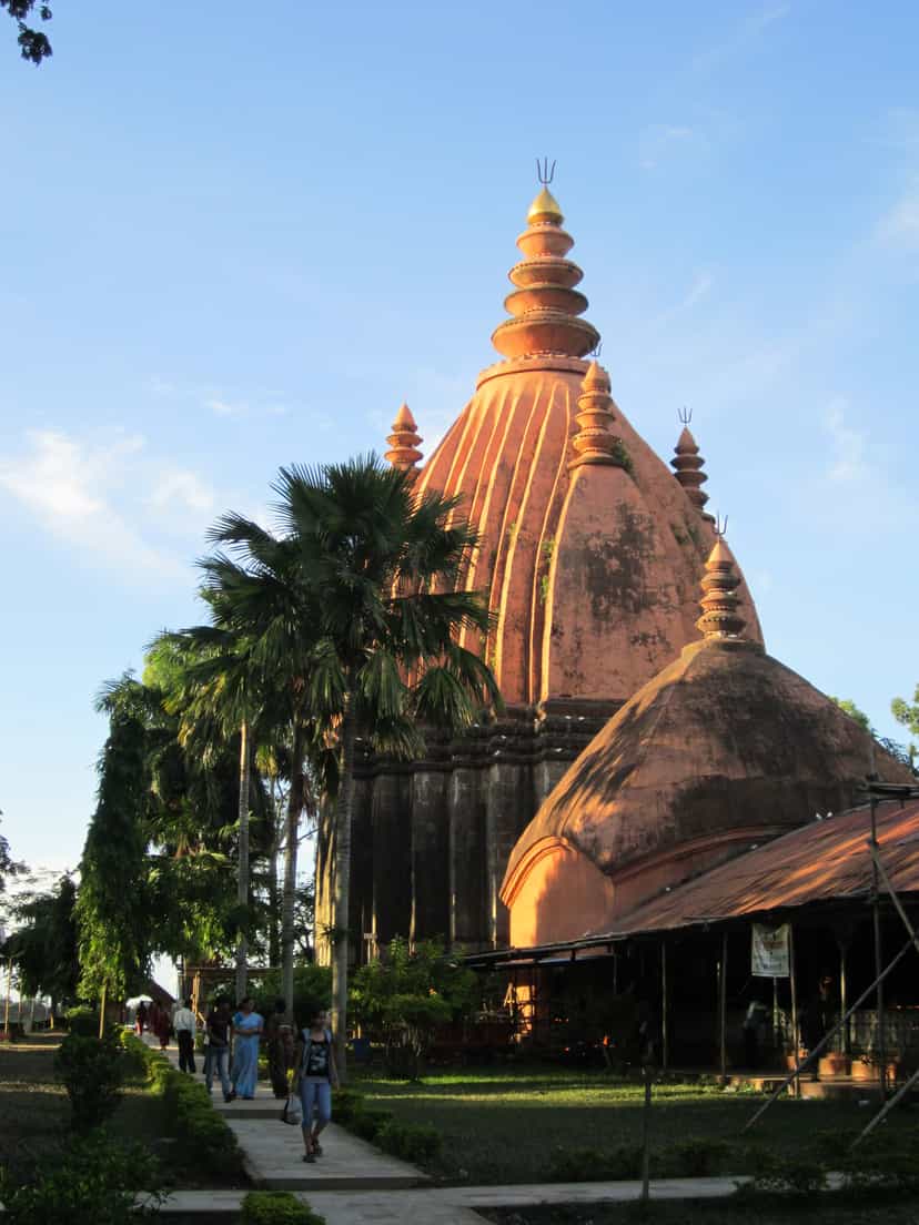 The towering red-ochre brick spire of the Sivadol at Sivasagar glowing in late-afternoon golden light against a deep blue sky, framed by a tall palm