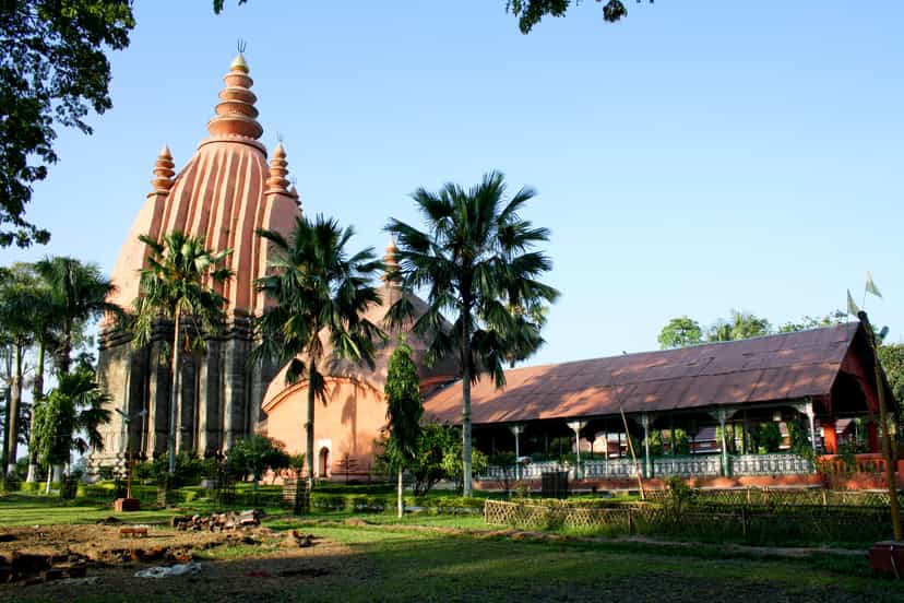 Wide view of the Sivadol temple group at Sivasagar in warm light: the tall brick spire and a companion domed shrine rising behind a long pitched-roof mandapa hall, with palms and a garden in front