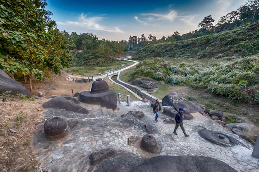 Rounded votive stupas cut from dark granite boulders on a cleared slope, a white stone path winding down into a green valley under a golden evening sky