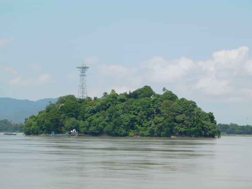 The wholly wooded Peacock Island rising from the broad Brahmaputra under a blue sky, the ferry landing at its near shore and low hills along the far bank
