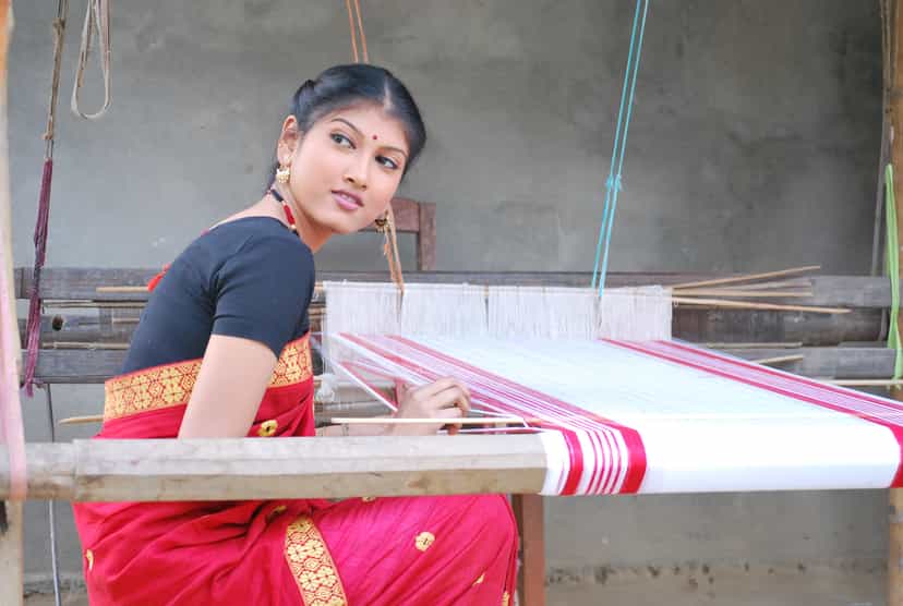 A woman in a traditional Assamese mekhela sador seated at a wooden handloom, a red-and-white gamosa taking shape in the warp before her