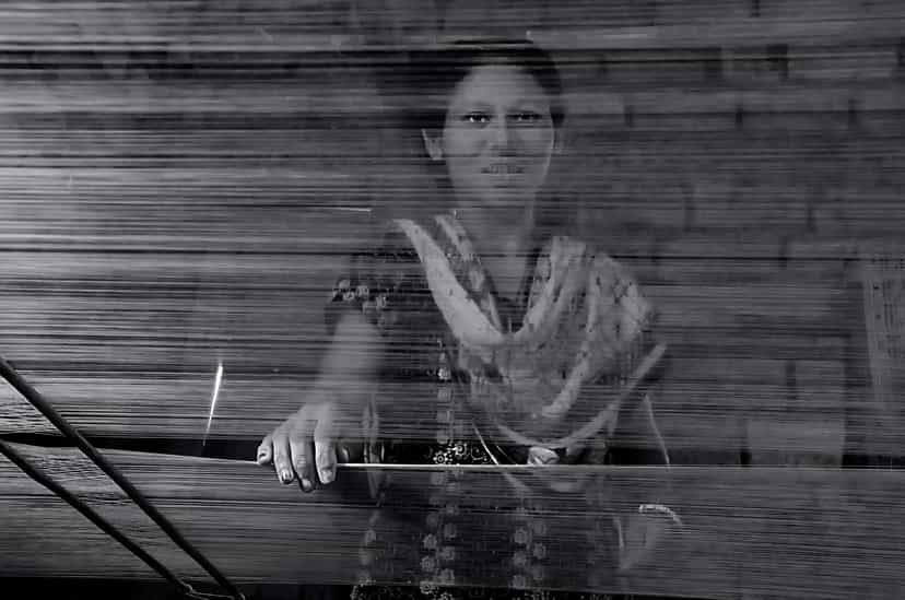 A weaver seen through the stretched warp threads of a handloom, her hand resting on the loom beam, in Sualkuchi