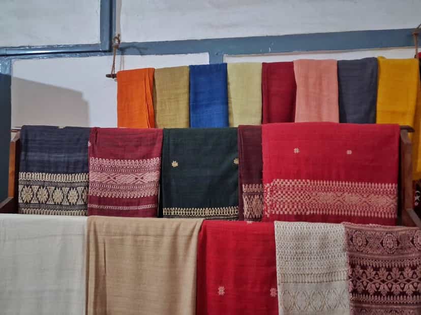 Rows of handwoven Assamese silk stoles and shawls hung on a wooden rack, in muga gold, eri cream and dyed red, blue, green and orange, each with woven floral and geometric border bands