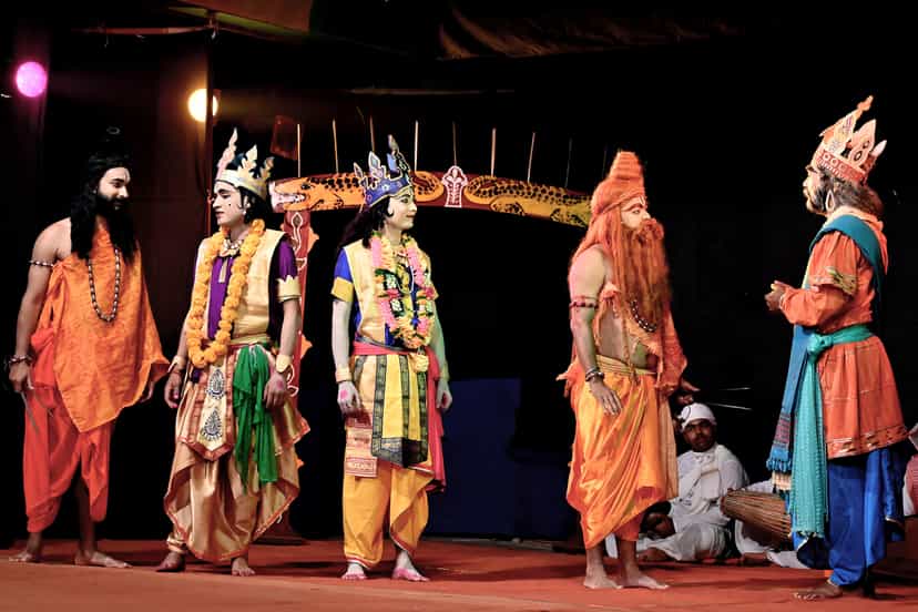 Costumed performers in robes, garlands and crowns stand on a sattra stage with a khol drummer seated behind them