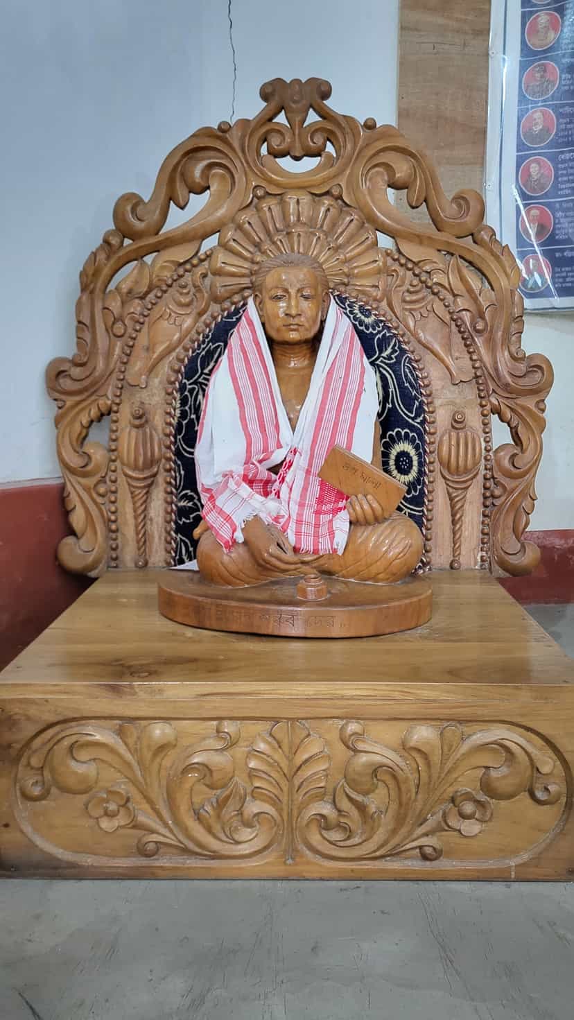 Carved wooden statue of Srimanta Sankardev seated cross-legged on a lotus pedestal within an ornate wooden halo-frame, draped in a red-and-white Assamese gamosa and holding a small inscribed manuscript