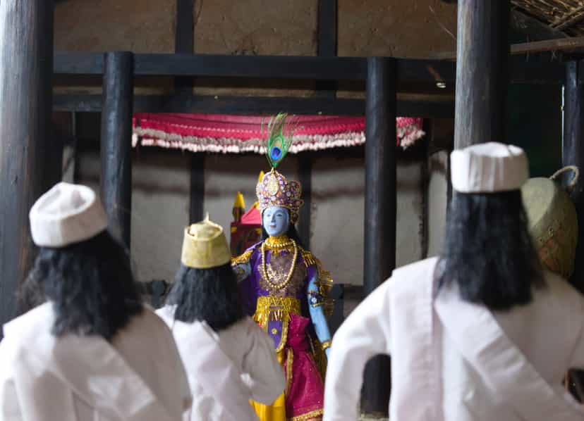A bhaona scene: a blue-painted figure of Krishna in a jewelled peacock-feather crown and purple-and-gold robe stands beneath a red canopy in a thatched namghar hall, with white-clad gayan performers in white caps seen from behind in the foreground and a khol drum at the right