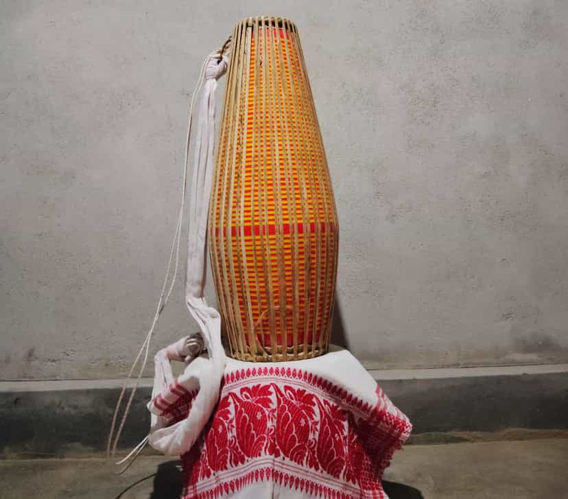 A single two-headed khol drum standing upright on its narrower head, its long tapering body wrapped in close vertical bamboo-and-cane lacing over orange and red banding, a white cloth carrying strap looped down one side, the drum resting on a folded red-and-white gamosa against a plain grey wall
