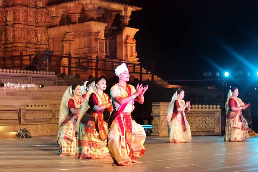 A group of Sattriya dancers in red and gold silk costumes, the central male dancer wearing the white cloth turban, performing on a lit stage before a temple