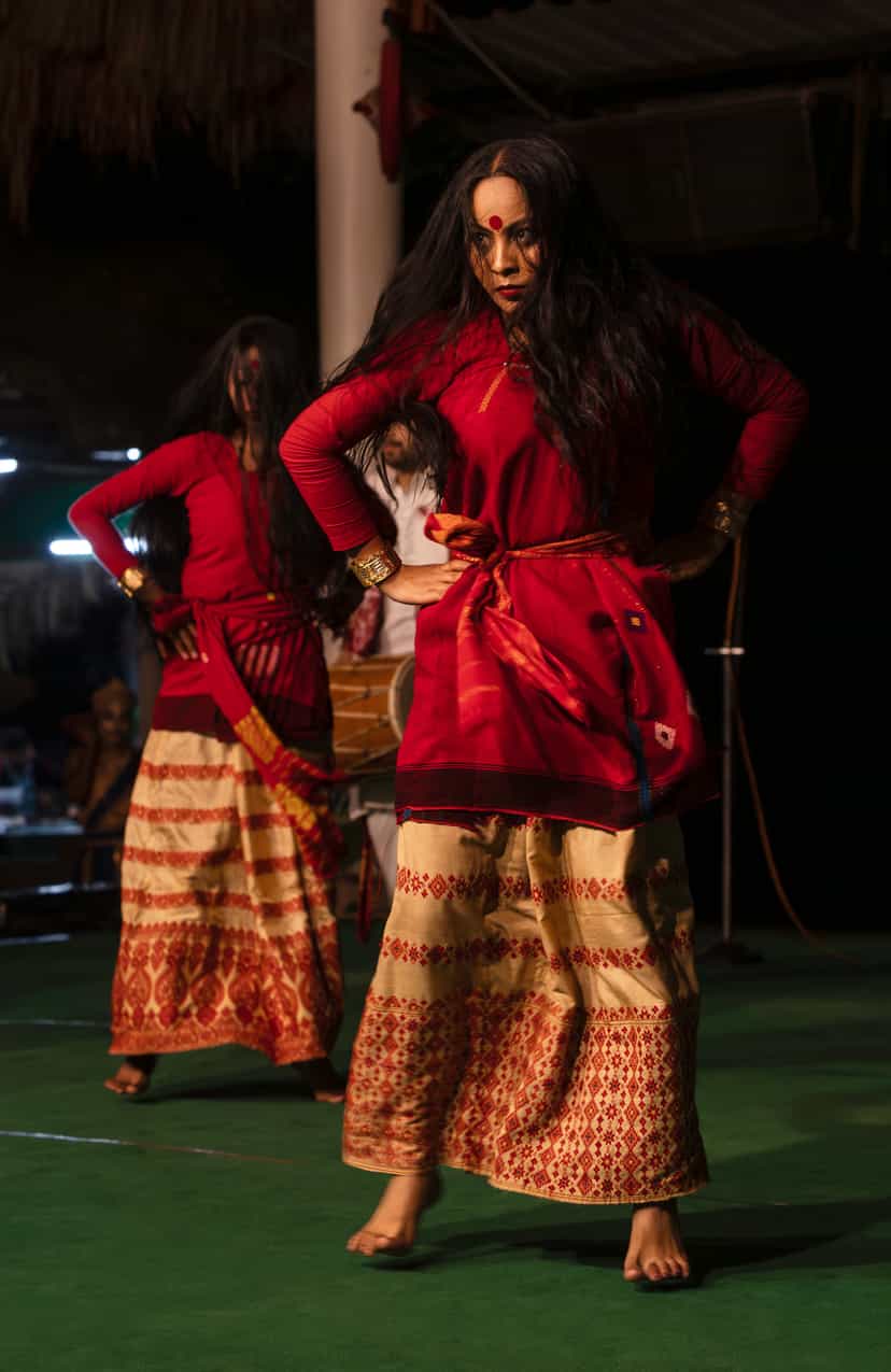 A Sattriya dancer in a red blouse and cream Muga silk skirt woven with traditional Assamese motifs, gold cuff bracelets, a khol drum visible behind