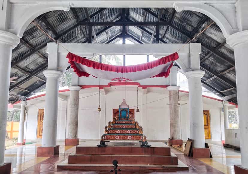 Interior of a Vaishnavite namghar prayer hall with white pillars and a central decorated altar throne flanked by hanging bells