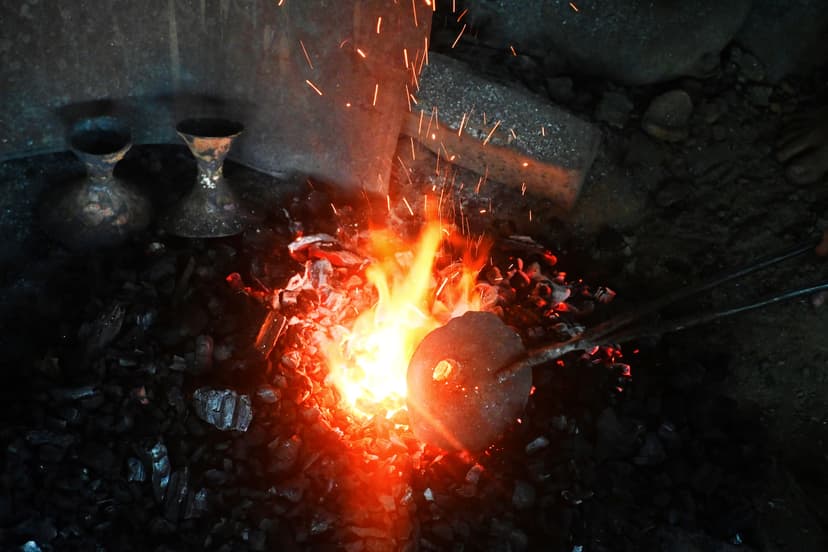 A rounded lump of bell-metal held on an iron rod glows in a bed of burning charcoal, sparks rising; two finished bell-metal vessels sit in the dark at the back of the forge