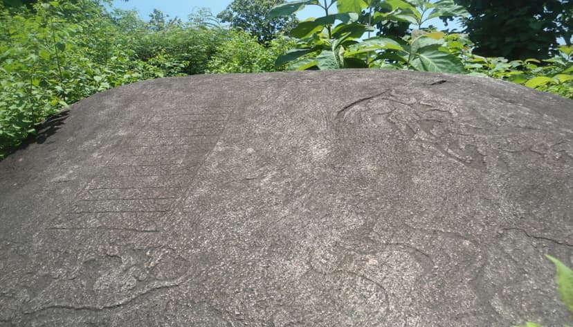 Weathered grey boulder near Tezpur carved with lines of Assamese script and flanking motifs, set among green foliage under a clear sky