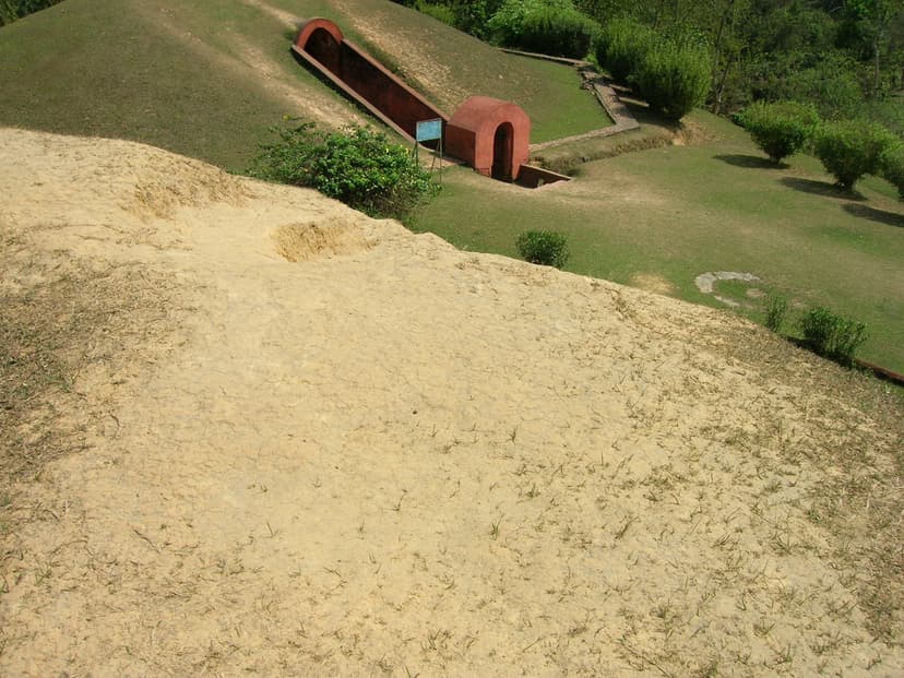 Looking down the grassy crown of one earthen burial mound toward the red brick arched vault entrance of an adjacent maidam, set into a mowed lawn with shrubs and a small signboard