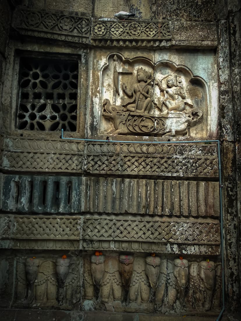 A weathered stone temple wall: a lobed niche holds a relief of two figures seated in a chariot, beside a pierced lattice window, above bands of geometric carving and a basal row of carved elephants with marigold offerings