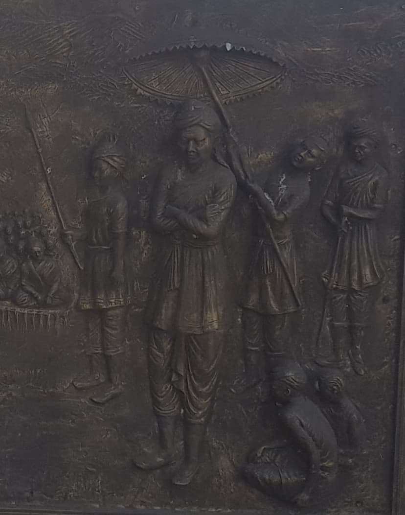 Bronze relief panel of Momai Tamuli Borbarua, the first Borbarua, standing beneath a ceremonial parasol with attendants