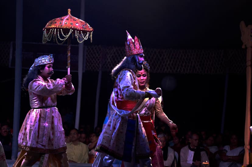 Costumed players in a night bhaona, a crowned figure under a decorated umbrella, the audience seated on the floor