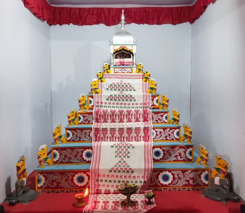 The seven-tiered wooden guru-asana of an Assamese namghar, painted red, blue and white and flanked by carved yellow lion figures on each tier, a silver canopied throne at the top holding the sacred book, a long white gamosa printed with the names Hari, Ram and Krishna in Assamese script running down the steps, a lit earthen lamp and an offering tray at its foot