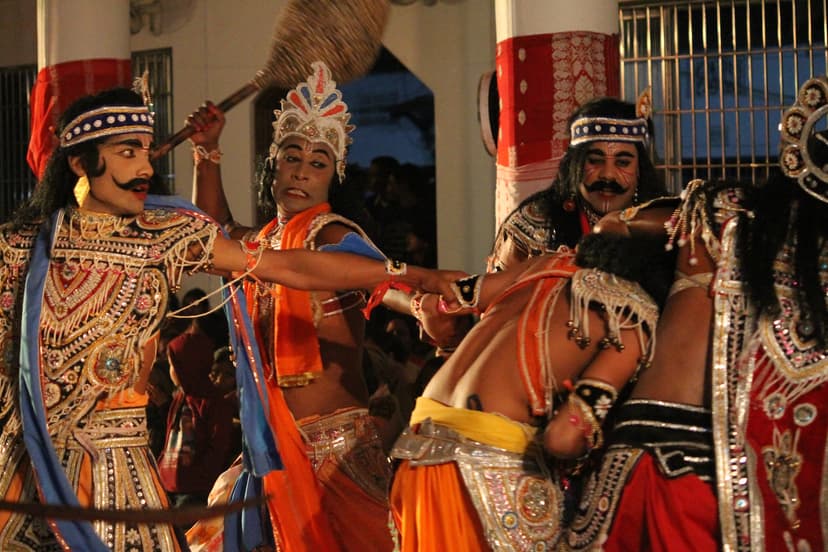 Four bhaona actors in heavy spangled costume mid-scene at night, faces painted with drawn moustaches and brows, wearing jewelled crowns and headbands, one raising a mace, another gripping a rival's wrist, before a pillared hall hung with a red and white cloth