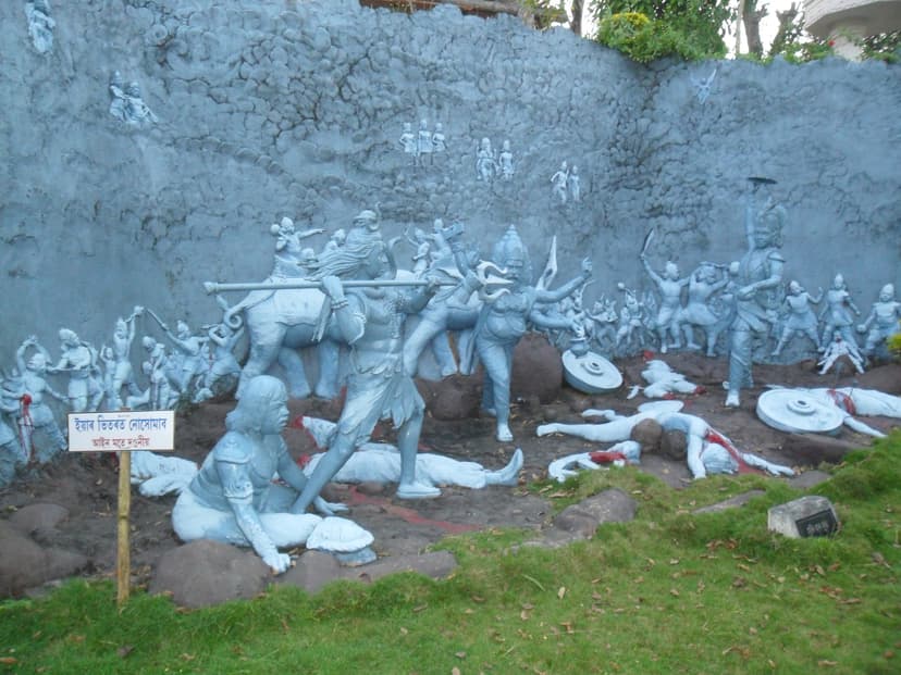 A large grey sculptural tableau against a rock wall: Shiva with raised trident and Krishna on his mount above ranks of fighting figures, with fallen warriors and shields strewn on the ground below, red paint marking their wounds