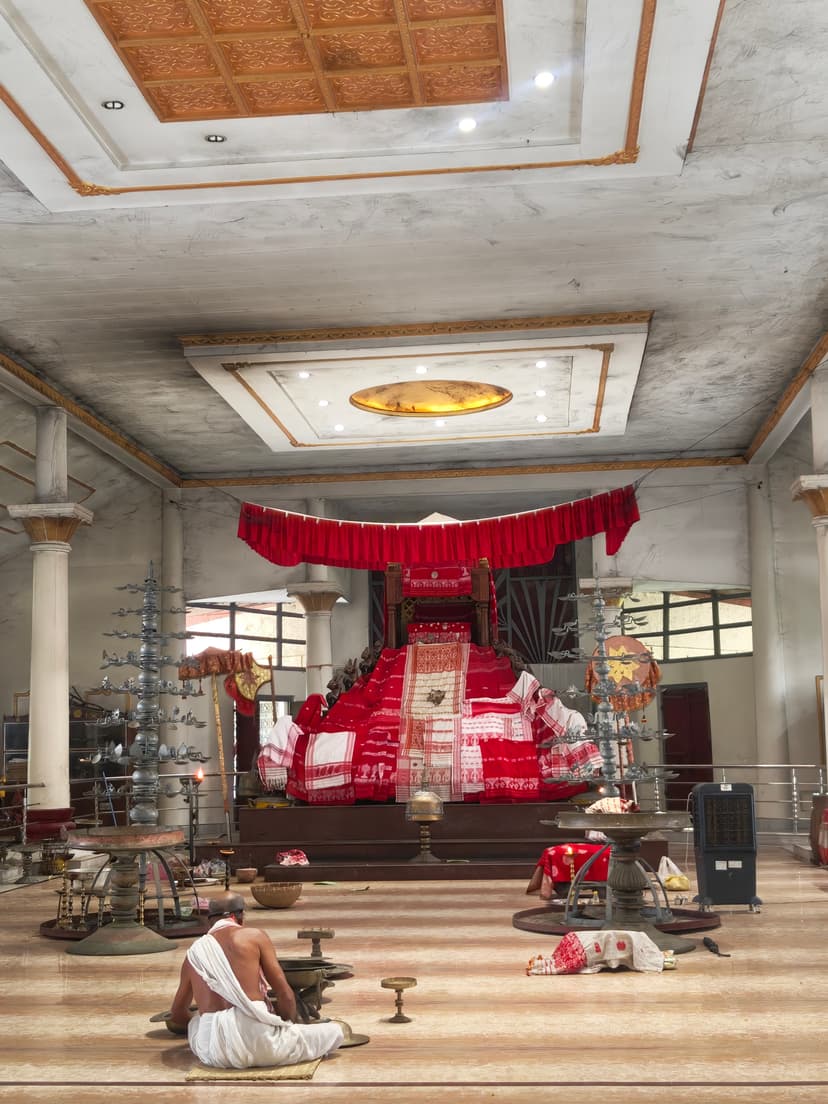 The marble-floored prayer hall of the Batadrava Than, with a red-draped throne bearing the sacred manuscript at its centre and brass ceremonial lamps to either side