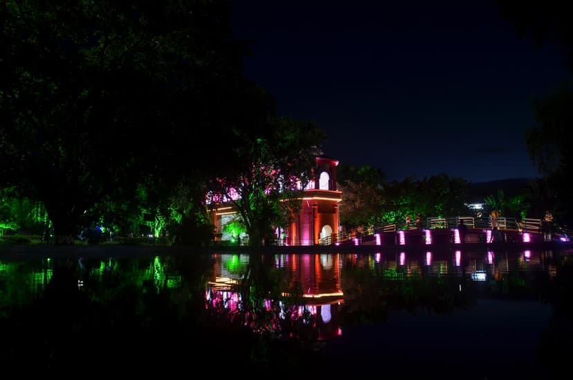 A lit octagonal brick pavilion and an arched footbridge glowing magenta reflect in a still pond at dusk, framed by dark trees